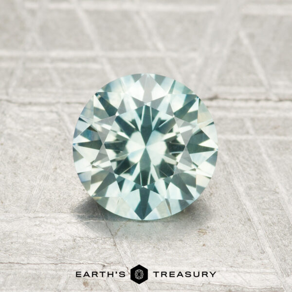 1.29-Carat Montana Sapphire (Heated) A 1.29-carat heated Montana sapphire with a round, faceted cut and pale blue-green hue is displayed on a textured light gray surface. "EARTH'S TREASURY" appears at the bottom.
