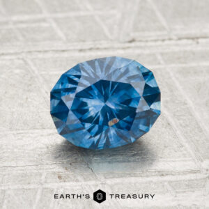 A round, faceted blue gemstone—1.49-carat heated Montana sapphire—rests on a light textured surface. "EARTH'S TREASURY" is displayed at the bottom of the image.