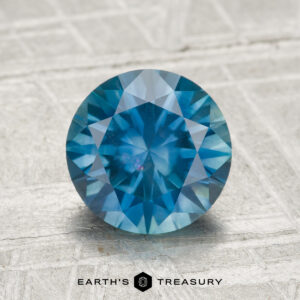A 2.04-carat heated Montana sapphire, round brilliant-cut, is displayed on a textured surface with "EARTH'S TREASURY" at the bottom.