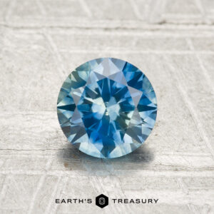 A 1.08-carat heated Montana sapphire, round and brilliant-cut, is displayed on a textured, light background with the "EARTH'S TREASURY" text and logo below it.