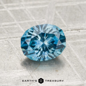 A 1.39-carat heated Montana sapphire with a faceted blue appearance rests on a textured silver-grey surface, with "Earth's Treasury" displayed at the bottom.