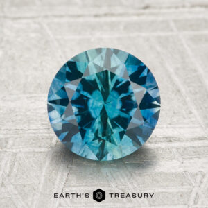 A 2.02-carat heated Montana Sapphire, round brilliant-cut and blue, displayed on a textured surface with the "EARTH'S TREASURY" logo at the bottom.