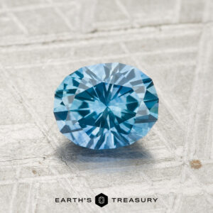 A 1.25-carat heated Montana sapphire is displayed on a light textured surface with the text "EARTH'S TREASURY" and a logo at the bottom.