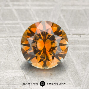 A 2.00-carat heated Montana Sapphire with round faceting and vibrant orange, brown, and yellow tones is displayed on a textured gray surface, with the "Earth's Treasury" logo beneath.