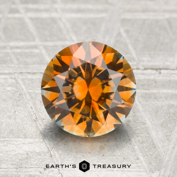 2.00-Carat Montana Sapphire (Heated) A 2.00-carat heated Montana Sapphire with round faceting and vibrant orange, brown, and yellow tones is displayed on a textured gray surface, with the "Earth's Treasury" logo beneath.