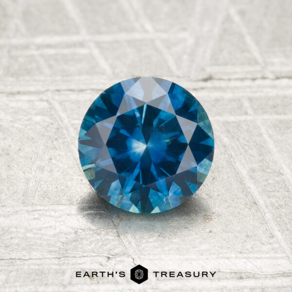 1.01-Carat Montana Sapphire (Heated) A 1.01-carat heated Montana sapphire with a round brilliant cut is showcased on a textured silvery surface, featuring the text "Earth's Treasury" at the bottom.