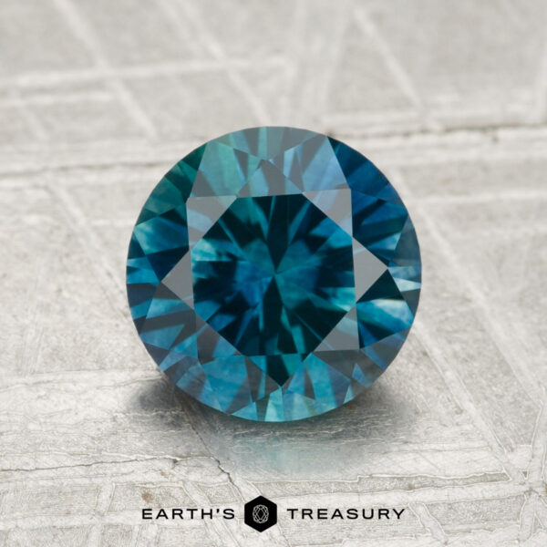 1.97-Carat Montana Sapphire (Heated) A 1.97-carat heated Montana sapphire, round brilliant-cut and teal in color, is displayed upright on a textured silver surface with "Earth's Treasury" at the bottom.