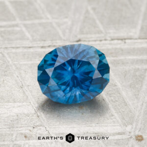 A 1.18-carat heated Montana sapphire, cut in a faceted oval shape, rests on a textured gray surface with “EARTH’S TREASURY” displayed at the bottom.