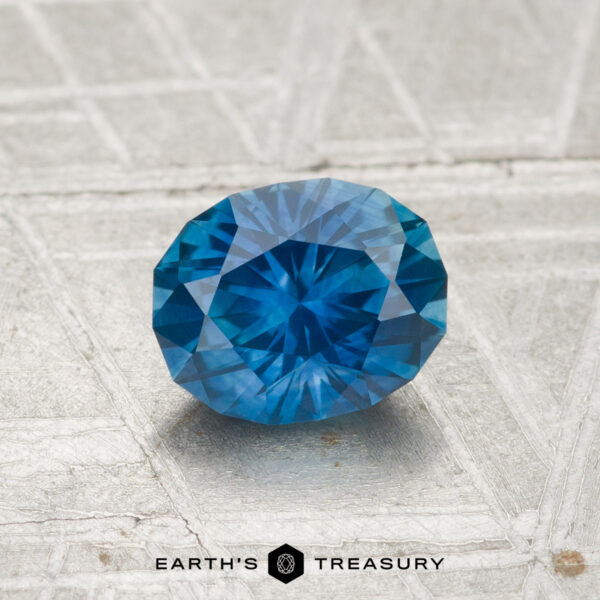 1.18-Carat Montana Sapphire (Heated) A 1.18-carat heated Montana sapphire, cut in a faceted oval shape, rests on a textured gray surface with “EARTH’S TREASURY” displayed at the bottom.