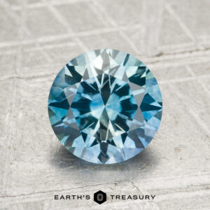 1.95-carat heated Montana Sapphire, round brilliant-cut with blue-green hues, shown on a textured surface featuring the "Earth's Treasury" logo at the bottom.