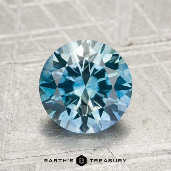 1.95-Carat Montana Sapphire (Heated) 1.95-carat heated Montana Sapphire, round brilliant-cut with blue-green hues, shown on a textured surface featuring the "Earth's Treasury" logo at the bottom.