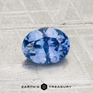 A 1.04-carat Ceylon Sapphire, faceted and oval-shaped, is displayed on a textured light gray surface with the "EARTH'S TREASURY" logo at the bottom.