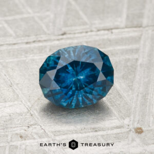 A 1.13-carat Montana Sapphire (heated), faceted in an oval cut and blue in color, is displayed on a textured, light-colored surface with "Earth's Treasury" beneath it.