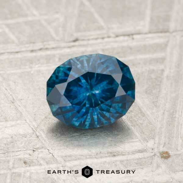 1.13-Carat Montana Sapphire (Heated) A 1.13-carat Montana Sapphire (heated), faceted in an oval cut and blue in color, is displayed on a textured, light-colored surface with "Earth's Treasury" beneath it.