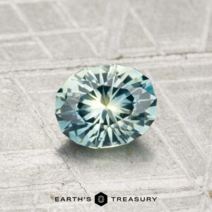 A round, faceted 1.01-carat Montana Sapphire (Heated) with blue-green hues rests on a textured gray surface. "EARTH'S TREASURY" text and logo appear at the bottom of the image.