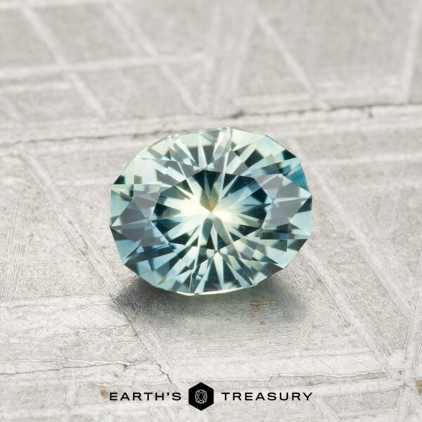 1.01-Carat Montana Sapphire (Heated) A round, faceted 1.01-carat Montana Sapphire (Heated) with blue-green hues rests on a textured gray surface. "EARTH'S TREASURY" text and logo appear at the bottom of the image.