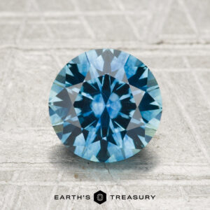 A round brilliant-cut 1.91-carat heated Montana sapphire is displayed on a textured gray surface, with the "EARTH'S TREASURY" text and logo at the bottom.