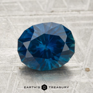 A 2.65-carat, oval-cut, deep blue Montana Sapphire (heated) rests on a light, patterned surface with "EARTH'S TREASURY" below.