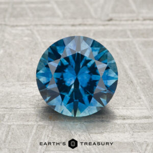 A 1.90-carat heated Montana sapphire in a round brilliant cut is displayed on a textured light gray background with "Earth's Treasury" at the bottom.