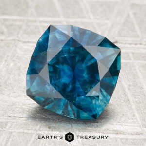 A 3.26-carat, cushion-cut Montana sapphire (heated) in deep blue, displayed on a textured light gray surface with "EARTH'S TREASURY" at the bottom.