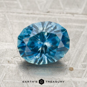 A 2.53-carat heated Montana sapphire with a brilliant cut sits on a patterned metallic surface; "EARTH'S TREASURY" appears at the bottom.
