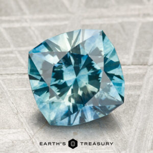 A 3.19-carat heated Montana sapphire, shimmering blue-green, rests on a textured surface with "Earth's Treasury" and its logo visible at the bottom of the image.
