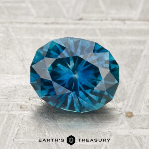 A 2.39-carat heated Montana sapphire, faceted in an oval shape, is displayed on a gray textured surface with the text "EARTH'S TREASURY" at the bottom.