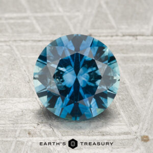 A 1.86-carat heated Montana sapphire, round brilliant-cut and blue in color, is displayed on a textured surface with "Earth's Treasury" visible at the bottom of the image.
