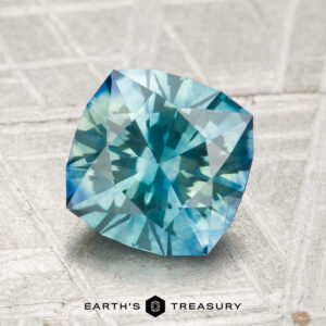 A 2.02-carat heated Montana sapphire with a cushion cut is shown on a textured surface, featuring the text "EARTH'S TREASURY" at the bottom.