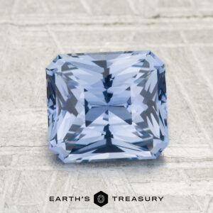 A 2.32-carat Ceylon sapphire, square-cut with intricate facets, rests on a textured gray surface. The "EARTH'S TREASURY" logo and text are featured at the bottom.