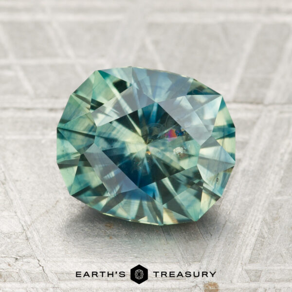 2.71-Carat Spruce Green-Blue ParticoloredMontana Sapphire (Heated) A 2.71-carat heated Montana sapphire with round, faceted cuts and blue-green hues rests on a textured, light surface. "EARTH'S TREASURY" is displayed at the bottom of the image.