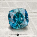 A 1.98-carat heated Montana sapphire with a cushion cut in teal blue is showcased on a textured light gray background, featuring the "Earth's Treasury" text below.