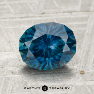 A 2.37-carat heated Montana sapphire with a faceted blue surface rests on textured light gray background, "EARTH'S TREASURY" shown below.