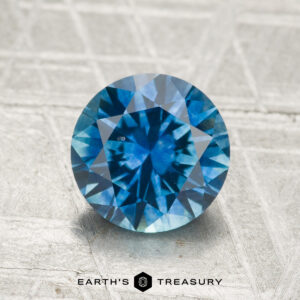 A 1.84-carat faceted Montana Sapphire (heated) with a blue hue is displayed on a textured light surface featuring the "Earth's Treasury" logo at the bottom.