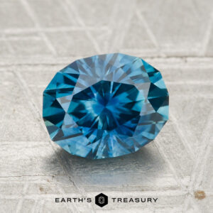 A 2.31-carat heated Montana sapphire with an oval cut and intricate facets is displayed on a textured light surface. The text "Earth's Treasury" appears at the bottom.