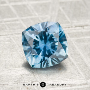 A 1.58-carat cushion-cut heated Montana sapphire with geometric facets is displayed on a textured gray surface, with the "EARTH'S TREASURY" logo and text below.