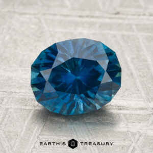 A 2.30-carat heated Montana sapphire, oval and blue, is displayed on a textured gray surface with "Earth's Treasury" written below.