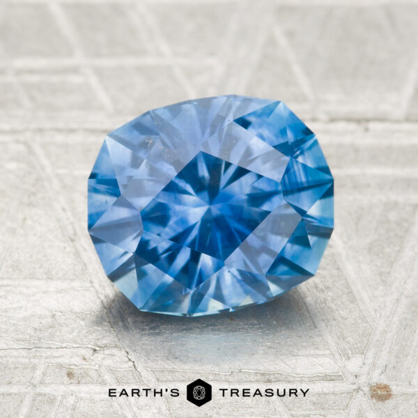 A 2.27-carat heated Montana sapphire, round cut and faceted in blue, rests on a textured gray surface with "EARTH'S TREASURY" displayed beneath it.