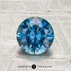 1.70-Carat Montana Sapphire (Heated), round brilliant-cut blue gemstone showcased on a textured light surface, with the Earth's Treasury logo featured below.