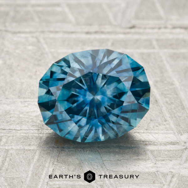 2.30-Carat Montana Sapphire (Heated) A 2.30-carat heated Montana sapphire, cut in a faceted oval shape, rests on a textured pale metallic surface with "EARTH'S TREASURY" displayed below.