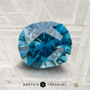 A 2.20-carat heated Montana sapphire with teal-blue hues rests on a textured surface; "EARTH'S TREASURY" text and logo are displayed at the bottom.