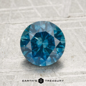 A 1.69-carat heated Montana sapphire in a round brilliant cut is showcased on a light textured surface with the "Earth's Treasury" logo displayed at the bottom.