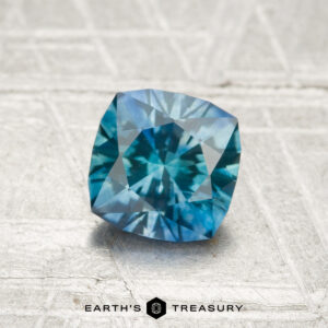 A 1.12-carat heated Montana sapphire with a cushion cut and blue-green hue rests on a textured grey surface featuring the "Earth's Treasury" logo and text at the bottom.
