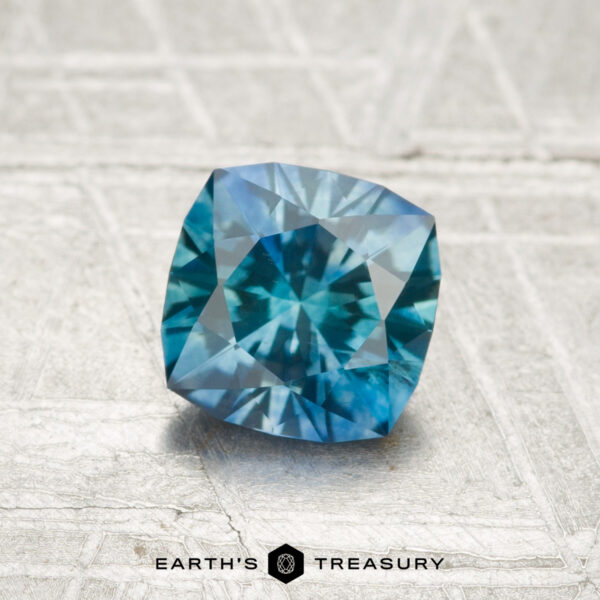 1.12-Carat Montana Sapphire (Heated) A 1.12-carat heated Montana sapphire with a cushion cut and blue-green hue rests on a textured grey surface featuring the "Earth's Treasury" logo and text at the bottom.
