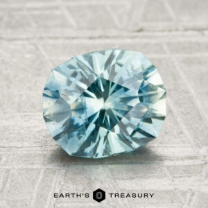 A faceted light blue 2.19-carat heated Montana sapphire is displayed on a textured surface with "Earth's Treasury" below it.