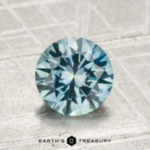 A round, faceted 1.68-carat heated Montana sapphire with a vibrant blue hue rests on a textured grey surface, with the "Earth's Treasury" logo and text visible at the bottom of the image.