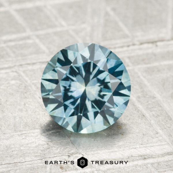A round, faceted 1.68-carat heated Montana sapphire with a vibrant blue hue rests on a textured grey surface, with the "Earth's Treasury" logo and text visible at the bottom of the image.