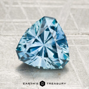 A 2.25-carat heated Montana sapphire, cut in a triangular shape and blue in color, is displayed on a textured light gray surface with the "Earth's Treasury" logo and text beneath it.