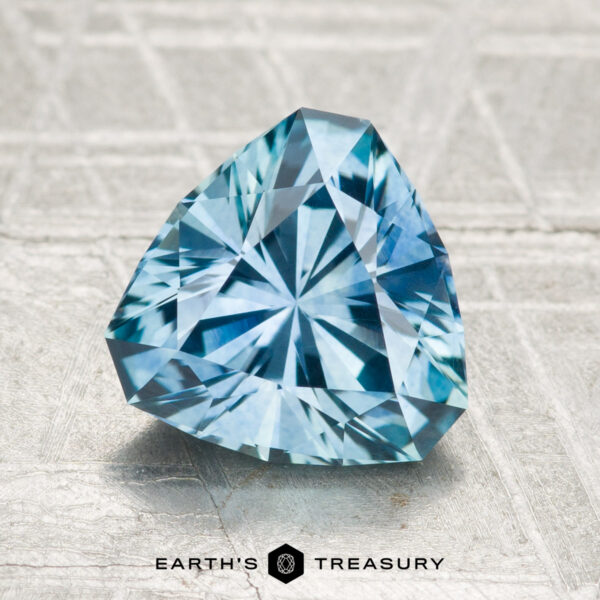 2.25-Carat Montana Sapphire (Heated) A 2.25-carat heated Montana sapphire, cut in a triangular shape and blue in color, is displayed on a textured light gray surface with the "Earth's Treasury" logo and text beneath it.