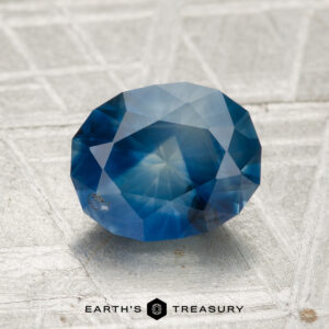 A 2.19-carat heated Montana sapphire, faceted in an oval shape, is displayed on a textured gray surface with the text "EARTH'S TREASURY" at the bottom.